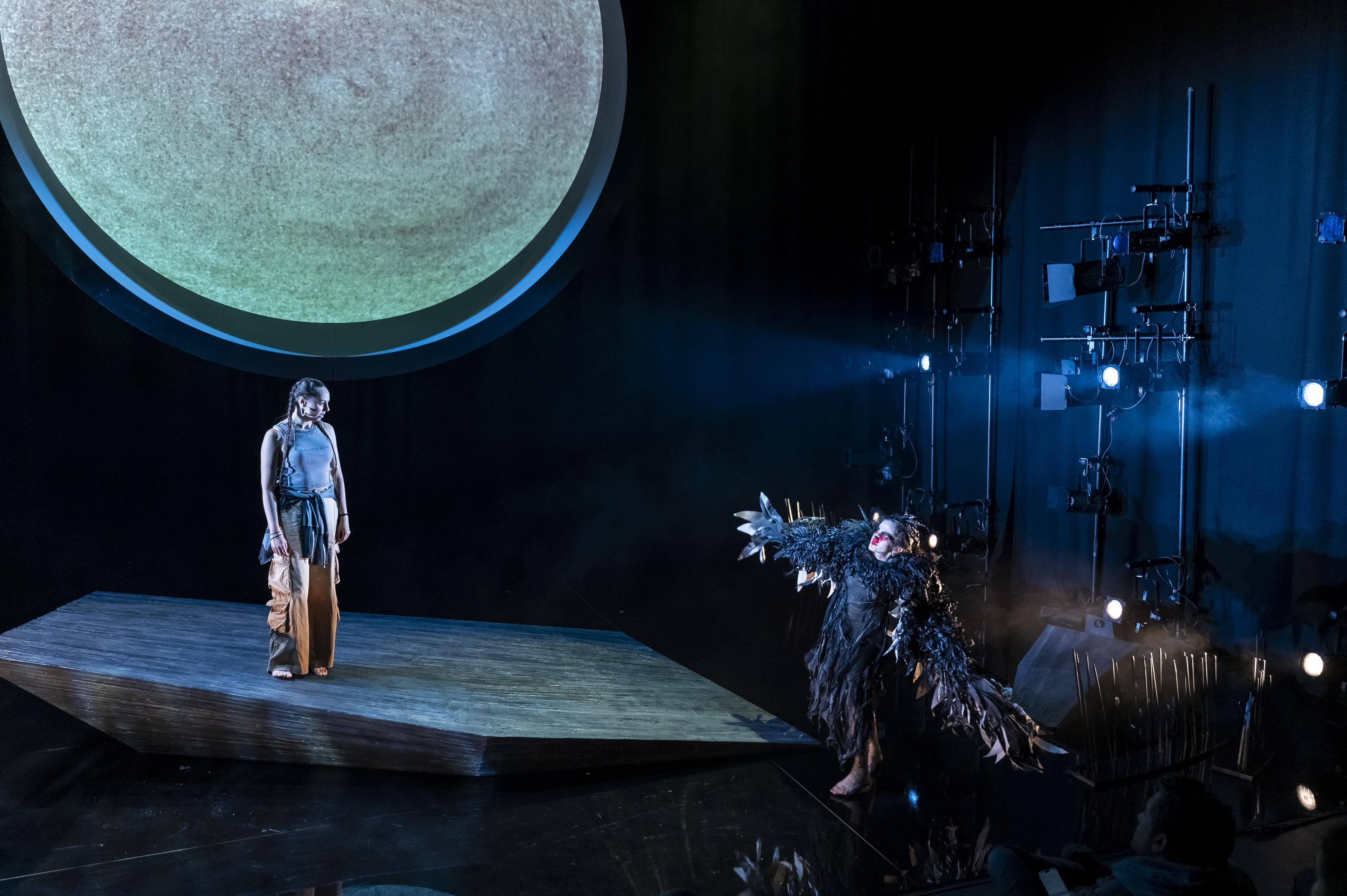 Actor performing on a dimly lit stage with a large moon shaped set design above them, looking down at another actor dressed as a bird in the MTC x ILBIJERRI production Blak in the Room: Gunnawarra Re-Creation.