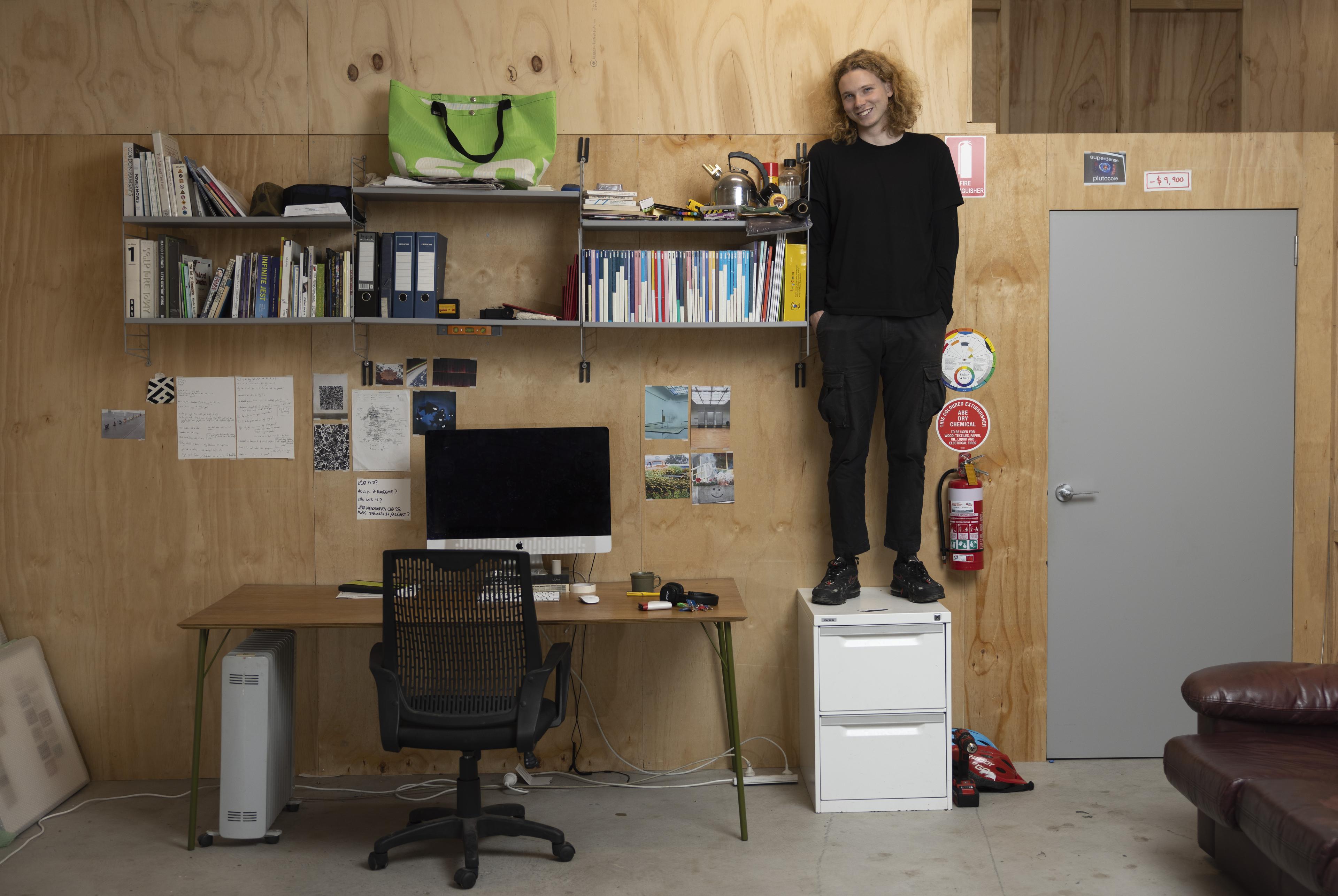A portrait of a man wearing all black standing on a small white filing cabinet in their studio, next to a desk with a computer on it, the wall behind them is all pine board.