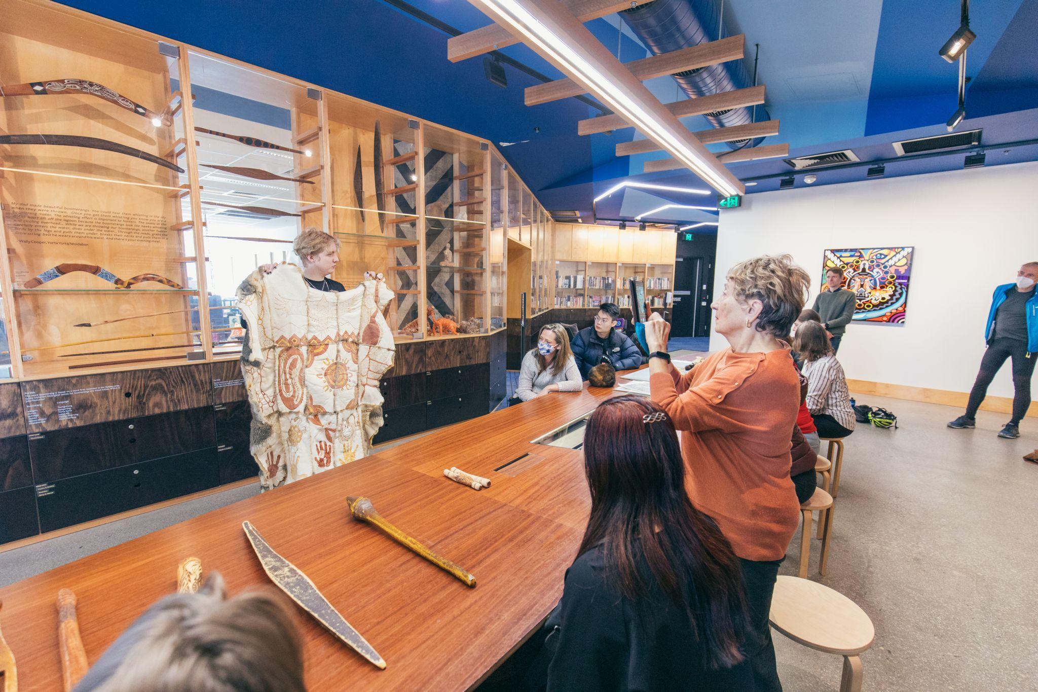 Visitors sitting around a large wooden table while a curator from Koorie Heritage Trust is standing up holding a possum skin coat and explaining the artwork on the coat.
