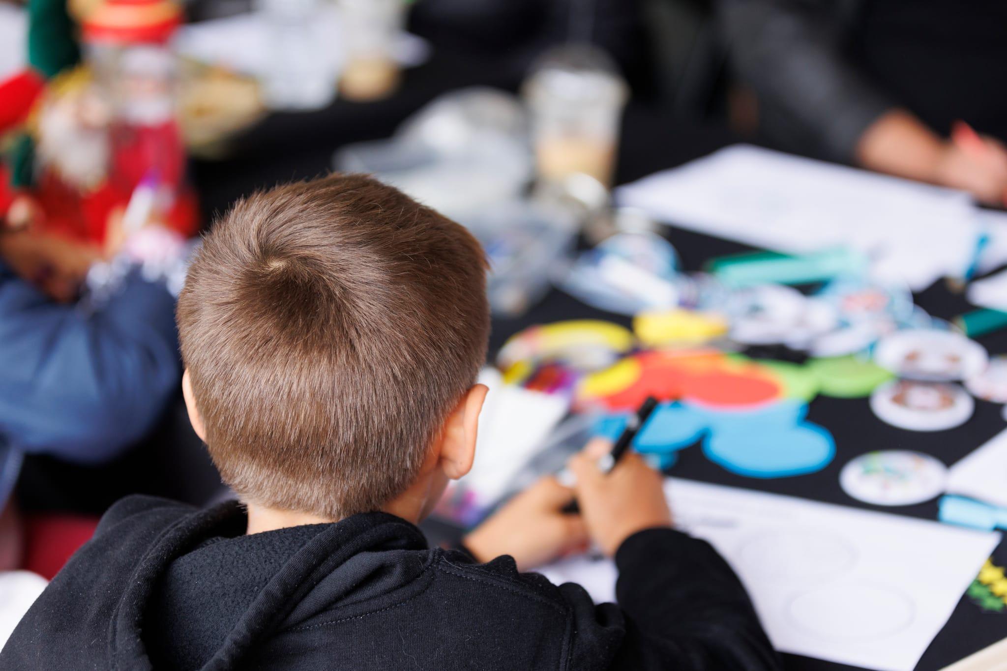 A child is facing away from the camera and can be seen drawing on paper with other arts and crafts items strewn across the table they are drawing on.