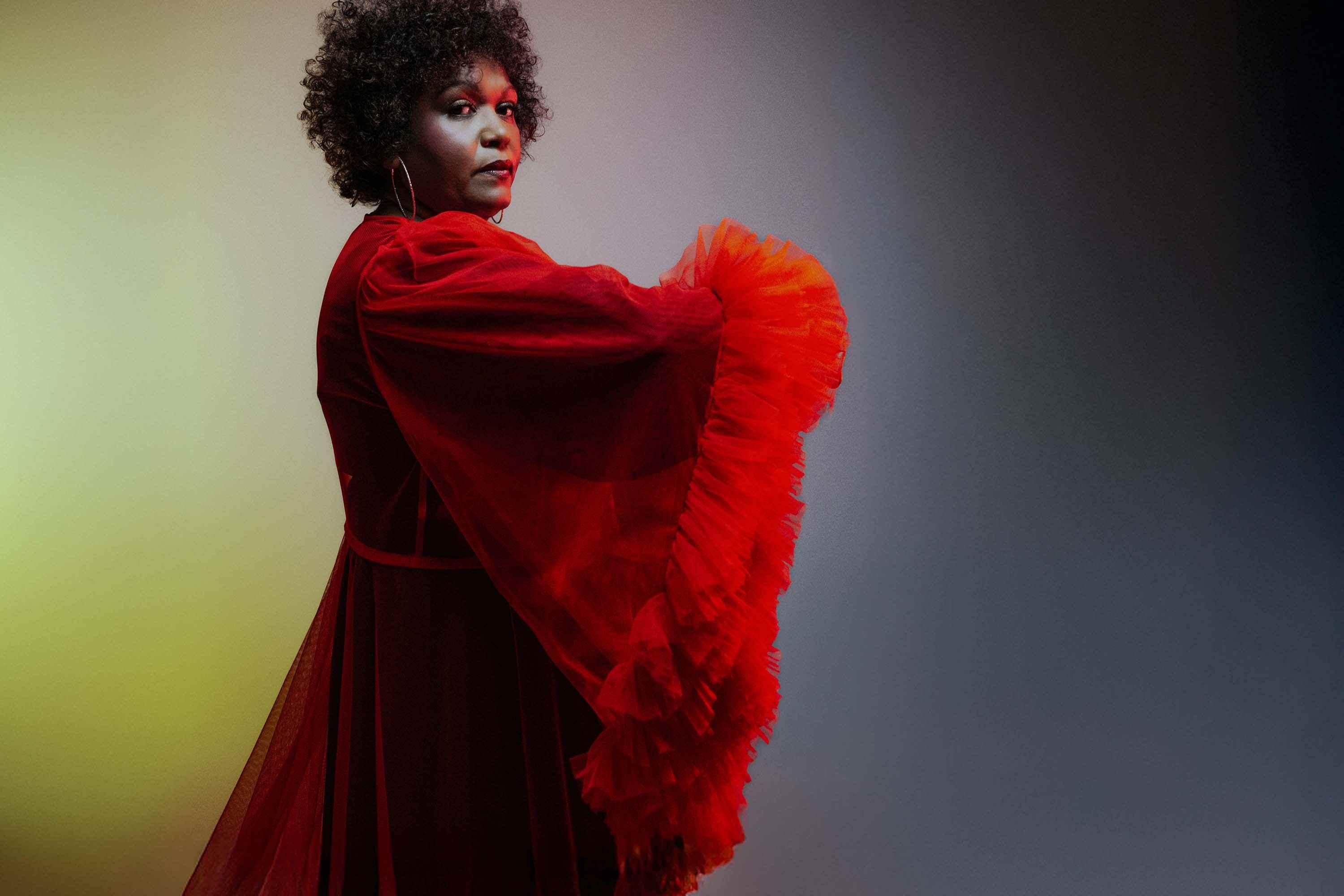 Singer Emma Donovan posing for a photo wearing a bright red dress in a profile position but looking at the camera.