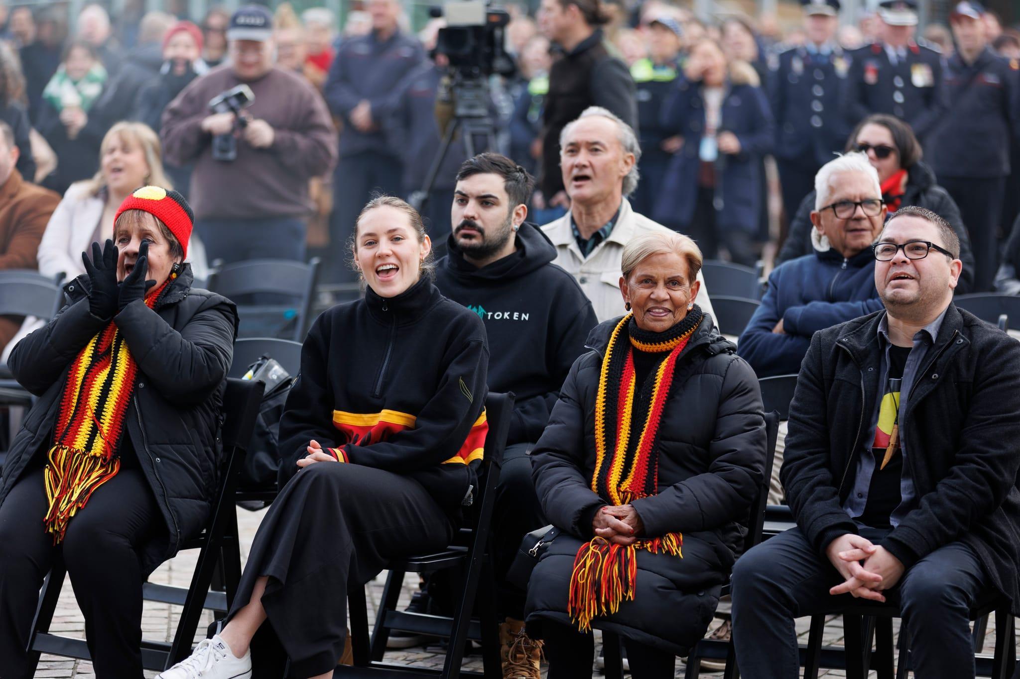 Crowd gathered outdoors sitting down and watching Aboriginal and Torres Strait Islander flags being raised on flagpoles during a NAIDOC Week ceremony.
