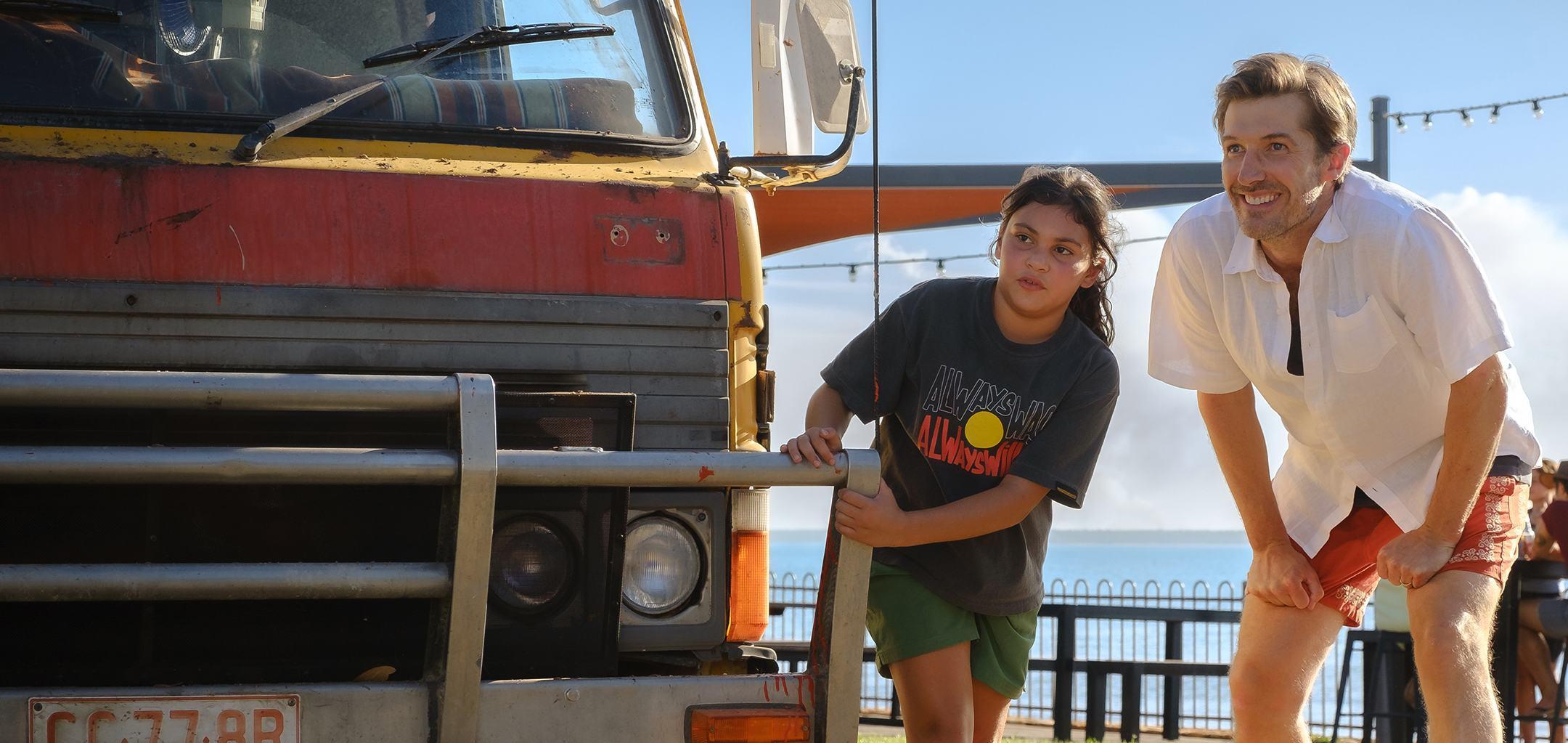 A young girl wearing an "Always Was, Always Will Be" t-shirt holding onto the bumper bar of a truck with an adult man in a white shirt crouching with his hands on his knees next to her. This is a promo photo from the Top End Bub film.