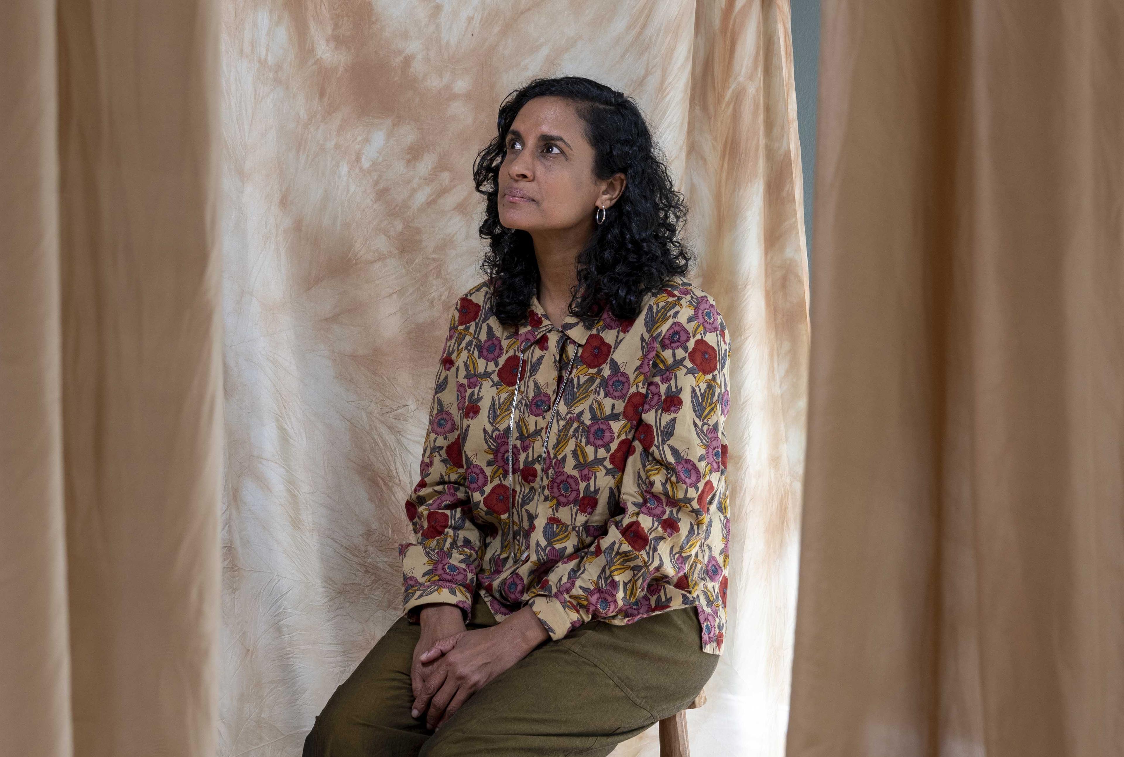 A portrait of artist Keg de Souza, she is sitting on a stool wearing a floral blouse, her hands are in her lap and she has a pensive look on her face looking up and away from the camera.