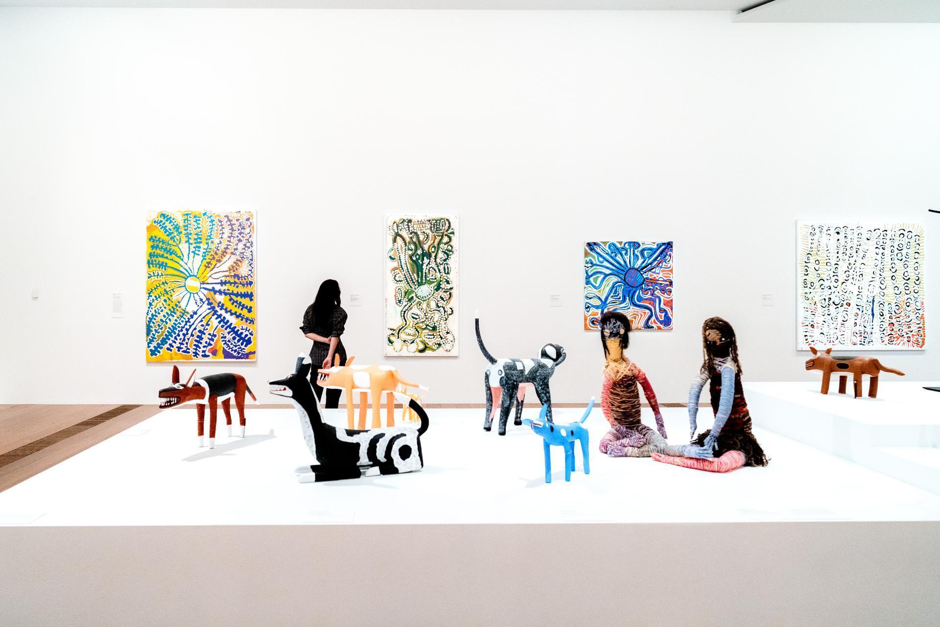 A person is viewing the Aboriginal gallery in NGV Australia there are colourful paintings on the walls and sculptures throughout the middle of the room.