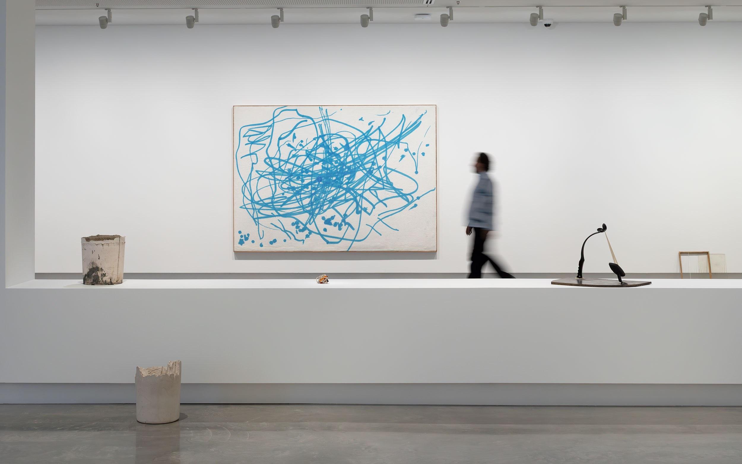 An image of a person walking trough a gallery, a large modern painting with blue squiggles all over it is hanging on the wall as the main focus.